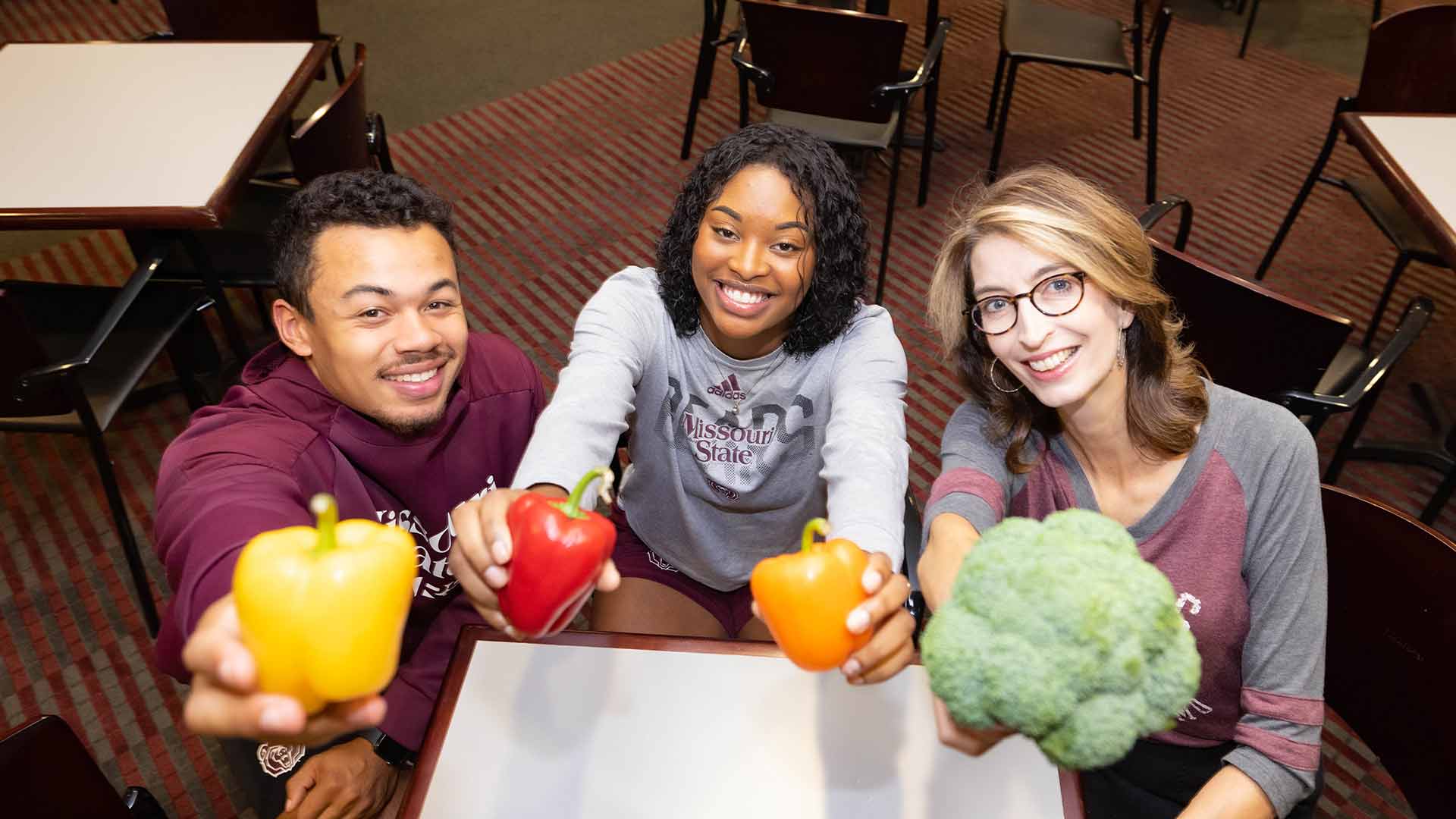 Two students and a professor smiling while holding up various vegetables close to the camera.